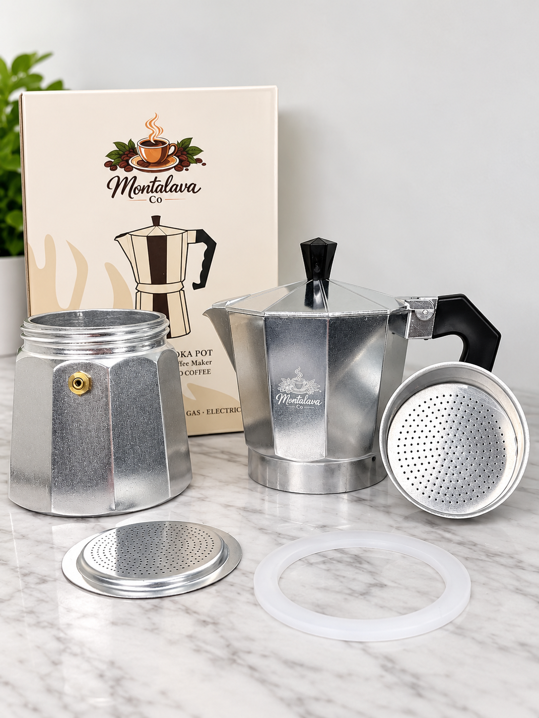 A silver aluminum moka pot set displayed on a white marble surface with its branded box behind it. The set includes the assembled stovetop coffee maker, a removed lid, a white silicone seal, and the product box featuring the Montalava Co logo.